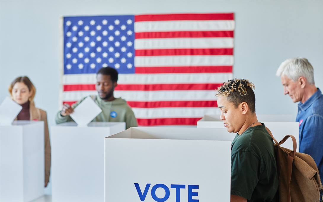 Several individuals at voting booths with an American flag in the background.