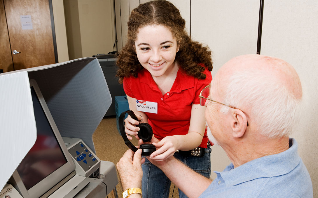 A smiling young woman with curly brown hair and a nametag that reads "Volunteer". The woman hands an older gentleman who has fair skin, glasses, and white hair a pair of headphones so that he may use the Voter Assist Terminal (VAT) to vote.